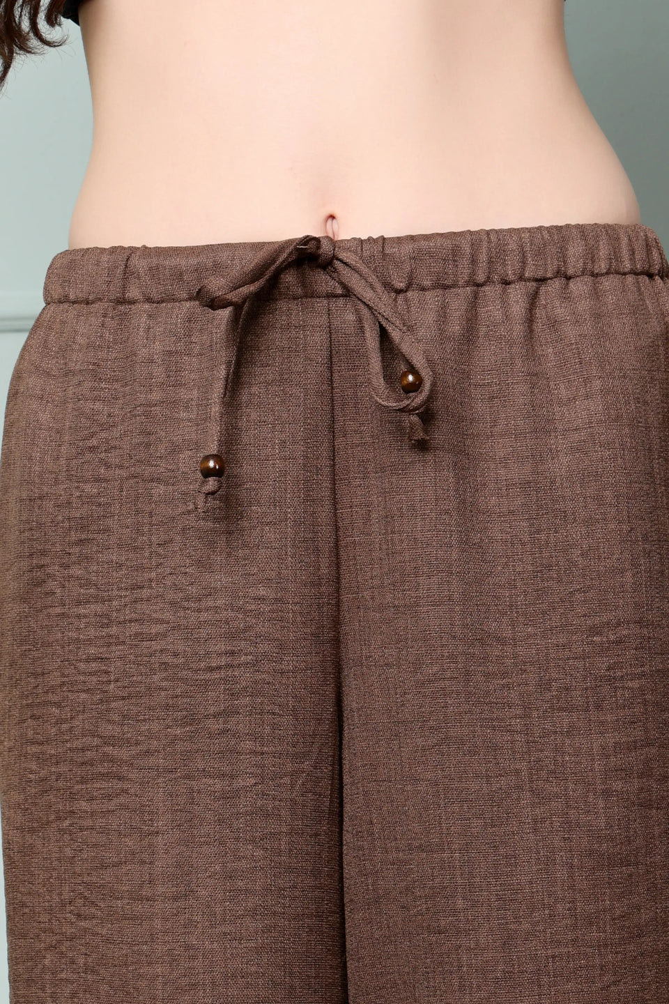 Close-up of RABRIC chocolate brown linen trousers with elastic waist, drawstring and wooden ring detailing