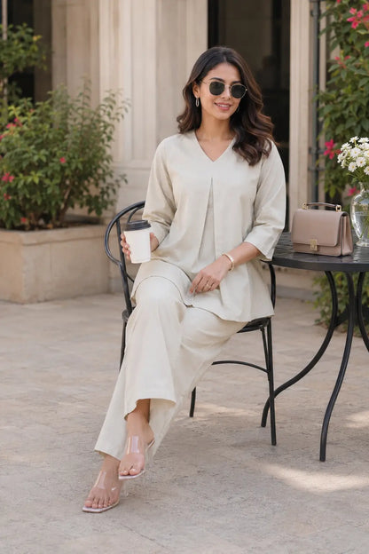 Rabric light beige cotton co-ord set for women in cafe sitting pose with V-neck pleated top, wide pajama and casual accessories